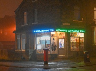Photograph of a brick building that is Broadways Convivence Store, blue and green LED lights display the shop sign. 