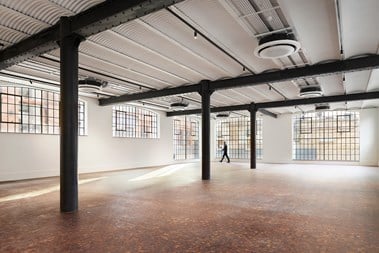 An interior photograph of an empty open-plan office space with black girders, white walls, and large, latticed windows. A person is walking around the room in the background.
