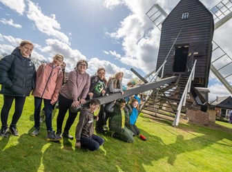 A group of young people stand next to the post that turns the Bourn Windmill. The mill can be seen to the right.