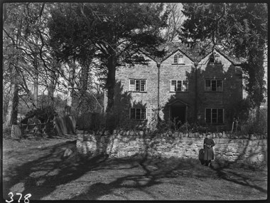 Black and white photograph of a large, stone house set behind trees and cast in shadow. In front of the house is a stone wall and standing in front of the wall is a woman wearing a hat, jumper and skirt and holding a torch or lamp.