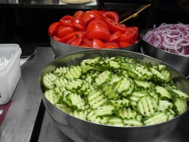 Large metal bowls full of sliced cucumber, tomoto and red onion.