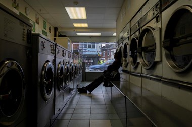 Interior view of a laundrette, with a person sitting on a bench silhouetted against the window waiting for their clothes. 