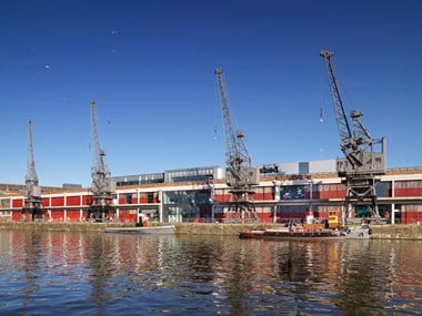 Four large, grey, metal cranes on a wharf in front of an industrial building.