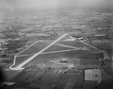 Black and white oblique aerial photograph of an airfield surrounded by fields. The three sections of runway are clearly visible and form a capital letter A shape on its side. A perimeter road skirts round part of the airfield. Just right of centre are the main airfield buildings, comprising several hangar-like structures. Beyond the airfield clusters of buildings and trees can be seen amongst the patchwork of fields. 