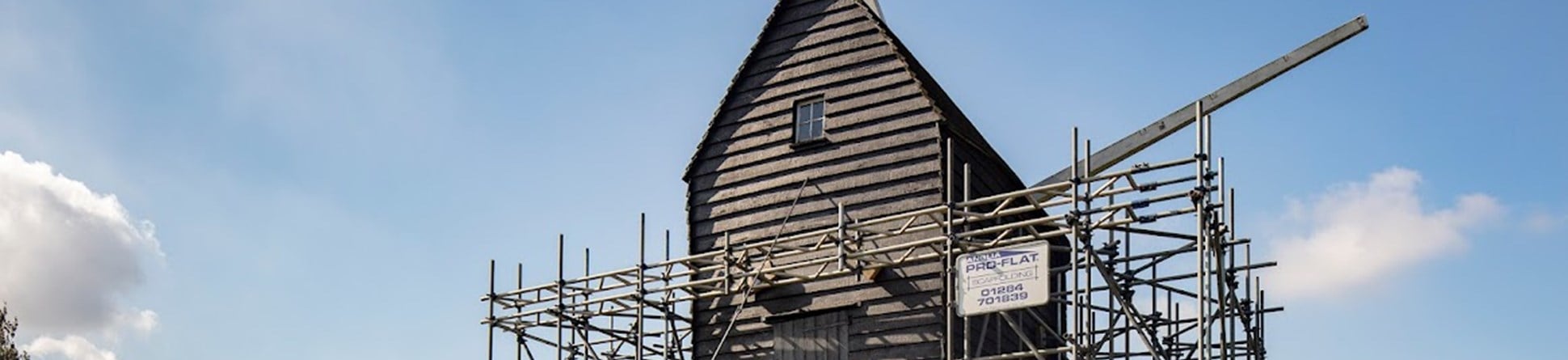 Bourn Windmill surrounded by scaffolding propping it  up. Steps are shown leading to a doorway. The post which turns the mill can be seen in the foreground. There's a small window towards the top of the mill and two sail posts can be seen on the other side of the building.