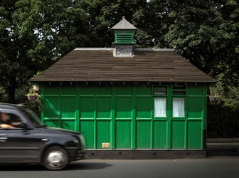 A small green wooden building with a small cote on the roadside with a modern black cab driving by.