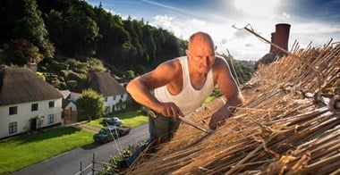 Close up photo of a man thatching a roof with other thatched houses visible in the background.