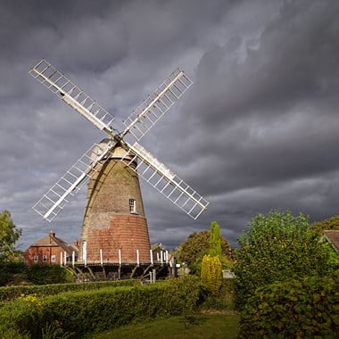 A windmill with white vanes but no sails a village setting stands in front of a stormy sky. 